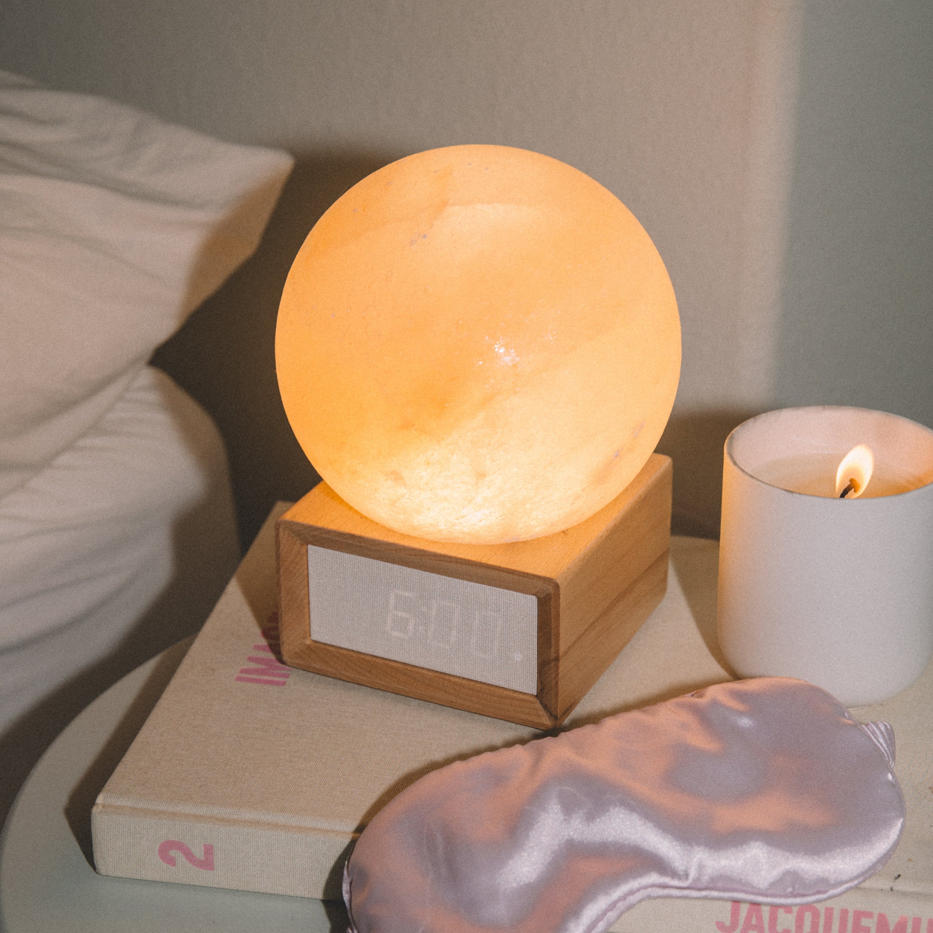 Himalayan salt sunrise alarm clock on a nightstand with a book, sleep mask, and candle.